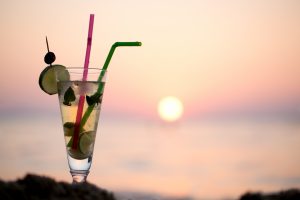 Close-up shot of glass with mojito cocktail with two straws on beach, sunset and sea in background